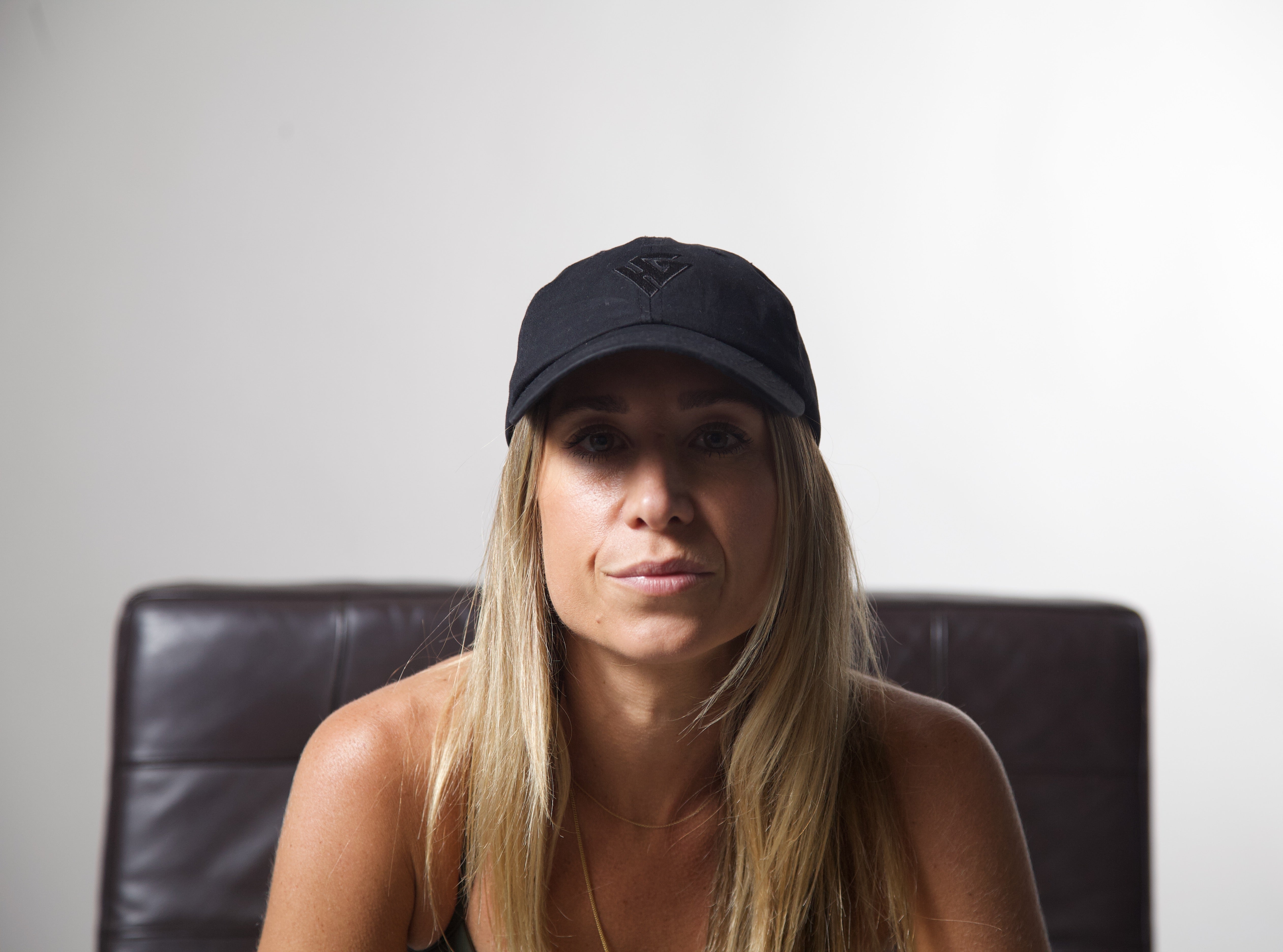 Woman wearing a black cap sitting on a brown chair against a plain white wall.